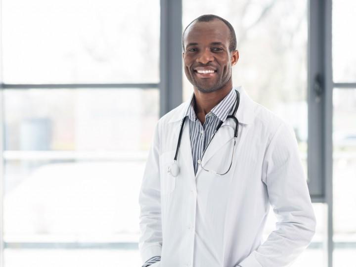 A doctor smiles and stands in front of window with white coat and stethoscope on